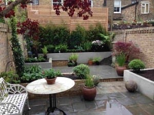 Small urban backyard with exotic potted plants, a circular table with a bench, slate paving, raised beds, and a wooden privacy screen against a brick wall.