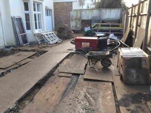 A construction site at the front of a building features concrete paths, wooden boards, a wheelbarrow, stacked materials, and various equipment.