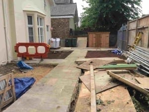 A paved pathway runs through a backyard under construction, with building materials, tools, and a wooden shed visible near the house’s front.
