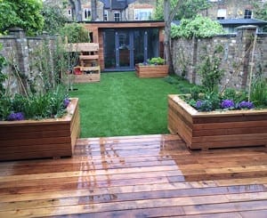 Wooden decking leads to a garden with artificial grass, raised wooden planters filled with green plants and flowers, and a stylish garden office or wooden shed at the back.