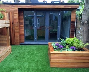 A modern wooden garden office room with large glass doors, artificial grass lawn, a wooden planter filled with flowers, and a wooden bench on the left.