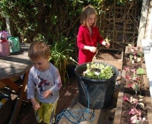 Two children are gardening; one is planting edible seedlings in a raised bed while the other stands nearby. There are plants, a bucket, and a watering can on a wooden patio.