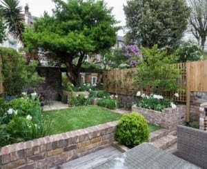 A small, neatly landscaped Brook Green backyard garden with brick walls, green lawn, flower beds, trees, and wooden fencing, featuring outdoor seating in the foreground.