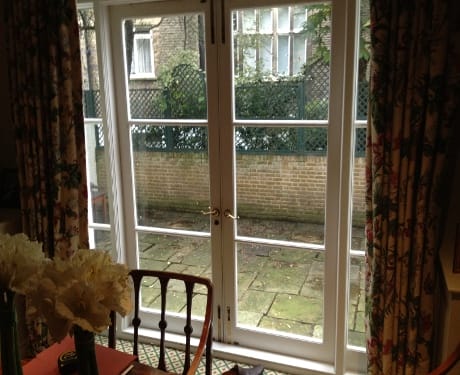 French doors with glass panes open to a small paved patio area outside. Patterned curtains, hinting at classic Knightsbridge elegance, frame the doorway, and a wooden chair is visible inside.