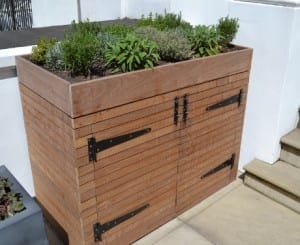 A wooden planter box ideal for container gardening, with herbs thriving on top, featuring double doors and black metal hinges, placed beside outdoor steps.