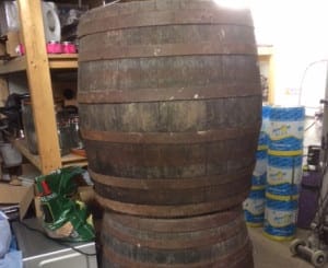Two large wooden barrels are stacked on top of each other in a cluttered storage area in Shepherd’s Bush, surrounded by shelves and assorted household items.
