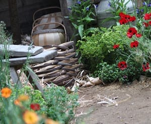 Wicker baskets are stacked near a wooden bench and surrounded by lush green plants and red flowers, creating a charming artisan gardens setting.