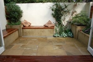 Small enclosed patio with stone flooring, built-in wooden bench seating, two patterned cushions, potted plants, and a white wall adorned with green foliage—perfect for inspiring small garden ideas and clever garden design solutions.