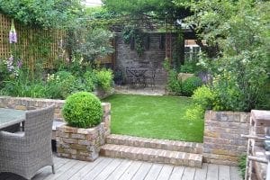A garden with artificial grass, raised brick planters, a patio area with wicker furniture, and a small metal table and chairs under a pergola with overhead beams, all surrounded by lush greenery.