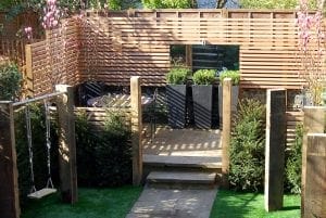 Enclosed garden with wooden fencing, patio seating area, potted plants, small trees, a rope swing, stone steps on artificial grass, and overhead beams ideal for vertical gardening.