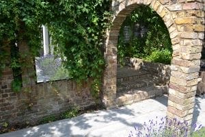 A brick archway leads to a small patio area surrounded by greenery and partially shaded by overhanging plants, with vertical gardening adding lush texture to the inviting space.