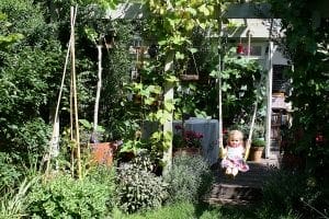 A young child sits on a swing under overhead beams of a leafy pergola, surrounded by lush green plants, flowers, and hints of vertical gardening in a garden that's worthy of the top 10 serene spots.