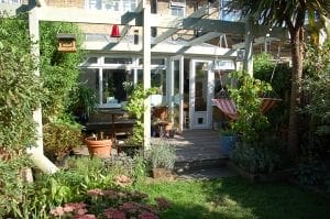 A backyard garden with a wooden deck, potted plants, vertical gardening accents, a pergola with overhead beams, outdoor furniture, and two striped hammocks near a glass door leading into the house.