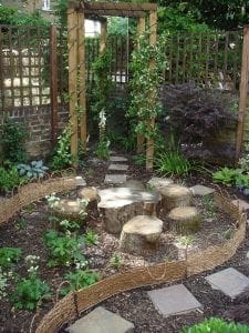 A small garden with stepping stones, wooden log seats, a woven fence, green plants, and vertical gardening features beneath a trellis arch covered in vines.