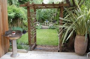 A backyard garden with a wooden pergola featuring overhead beams, a metal barbecue grill, potted plants, green lawn, and a stone bench in the background.