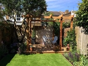 A wooden pergola with overhead beams shades a metal table and chairs on a deck, surrounded by lush plants, trees, and vertical gardening in a fenced backyard with artificial grass.