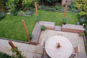A neatly maintained garden with a lawn, patio, overhead beams for vertical gardening, a round wooden table with four chairs, and raised flower beds featuring wooden posts and potted plants.