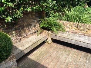 Wooden corner garden benches with brick supports surround a small fern planter, set against a brick wall and greenery in a sunlit outdoor patio area—an inviting spot for the best garden benches.