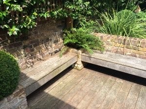 Wooden garden benches form an L shape around a corner with brick walls, surrounding a fern and other plants in a sunlit outdoor seating area.