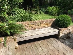 A wooden bench built into a low brick wall, surrounded by green plants and a manicured lawn, creates one of the best garden benches for relaxing on a wooden deck.