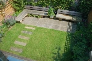 A small backyard garden with a grass lawn, stone path, rectangular stone patio, top benches for relaxing, and surrounding plants.