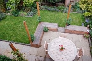 Aerial view of a backyard with garden furniture including a circular wooden table and chairs on the patio, top 10 benches, small plants, and a grassy lawn bordered by flower beds.