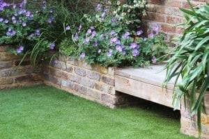 A brick raised flower bed with purple and white flowers and green plants sits beside one of the best garden benches, bordered by artificial grass.