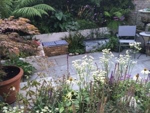 Small patio garden with flowering plants, a potted tree, and one of the best garden benches paired with a chair and table set against a brick wall, surrounded by dense greenery in the background.