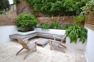 A small outdoor patio with two wooden chairs, a top bench, stone flooring, raised planters filled with green plants, and a wooden slat fence.
