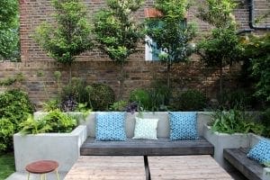 A modern garden seating area features stylish concrete garden benches with blue and white patterned cushions, wooden tables, and lush green plants set against a brick wall backdrop.