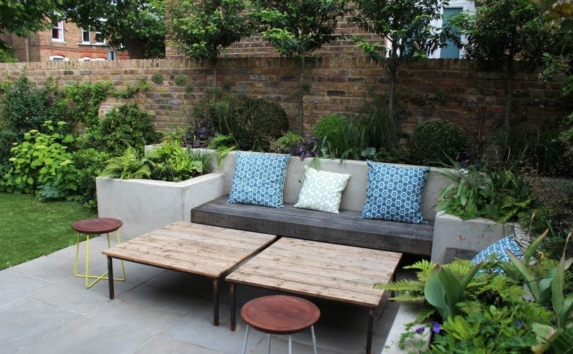 Outdoor patio area with stylish garden benches and a wooden sofa featuring patterned cushions, two wooden tables, and lush green plants and shrubs set against a charming brick wall.