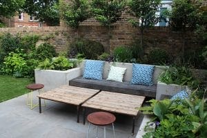 Modern patio with a wooden garden bench, patterned pillows, two wooden coffee tables, and surrounding greenery against a brick wall—showcasing one of the top 10 benches perfect for stylish gardens.