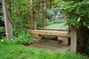 A wooden garden bench, perfect for outdoor seating, sits under a wooden trellis surrounded by green plants and grass.