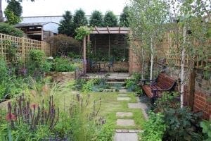 A landscaped garden with cottage style planting, a stone path, wooden bench, various flowering plants, and a pergola with a table and two chairs at the back.