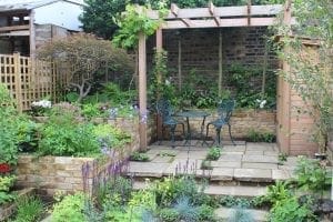 A small garden patio with two metal chairs and a table under a wooden pergola, surrounded by cottage style planting and brick walls.