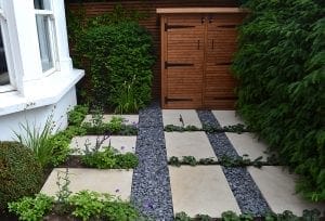 A small modern garden with large rectangular paving stones, gravel strips, green plants, and stylish bin sheds against a wooden fence.