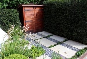 A wooden garden shed stands next to tall hedges, with a path of rectangular stone slabs bordered by gravel and green plants leading to this charming storage unit.