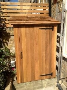 A small wooden storage unit with a sloped roof and a single door, situated against a brick wall next to a ladder.