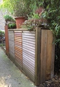 A wooden outdoor storage unit with horizontal slats, ideal for use as stylish bin sheds, topped with potted plants and flowers, positioned beside a path and surrounded by greenery.