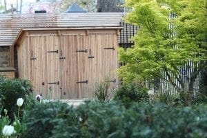 A wooden storage unit with three doors stands behind green bushes and a yellow-leaved tree in a garden. Tulips and other plants are visible in the foreground.