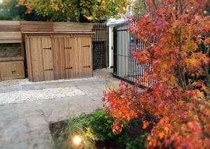 A landscaped garden area with a paved walkway, wooden bin sheds—perfect for top 10 storage solutions—a black metal gate, and an orange-leaved tree in the foreground.