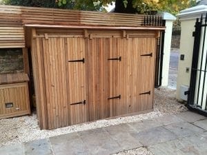 A wooden bin shed with double doors and black metal hinges stands on a gravel surface next to a paved walkway and a black metal fence, offering an attractive alternative to traditional storage units.
