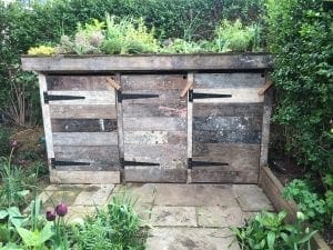 A three-bin wooden compost structure, reminiscent of stylish bin sheds, with a green roof planted with various small plants, situated on a stone patio in a garden.