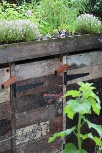 A close-up of a weathered wooden compost bin with metal hinges in a garden, topped with blooming herbs and flowers—an eco-friendly alternative to traditional bin sheds and storage units.