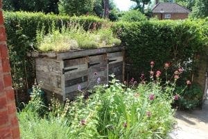 A weathered wooden beehive stands in a garden with flowering plants and greenery, nestled among bin sheds and storage units, with a tall hedge and a house in the background.