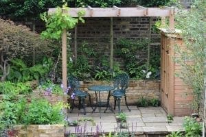 A small garden patio with a metal table and two chairs under a wooden pergola, surrounded by plants and flowers, next to storage units and a brick wall.