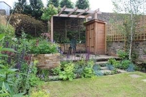 A small garden with flowering plants, a lawn, stone steps, a wooden shed, discreet bin sheds, and a pergola above a metal table and chairs near a brick wall.