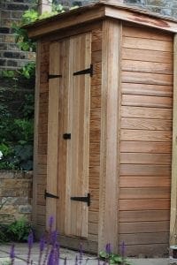 A small wooden bin shed with vertical planks, double doors, and black metal hinges stands beside a brick wall and some greenery.