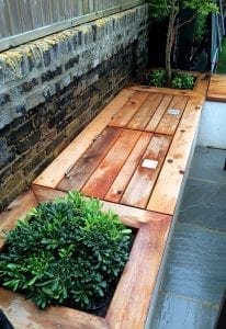 Wooden bench with built-in planters and a hinged storage compartment, offering the charm of outdoor storage units, set against a brick wall and slate tile flooring.