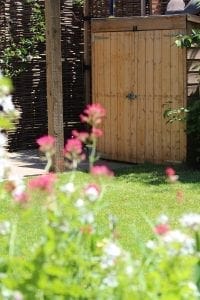 A wooden garden shed, ideal for use as storage units or bin sheds, stands with a closed door behind a green lawn and out-of-focus pink and white flowers in the foreground.